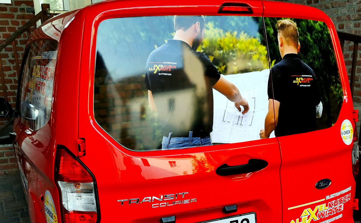 Zwei Elektriker von Kusch & Wirtz prüfen vor ihrem roten Firmenwagen einen Bauplan.